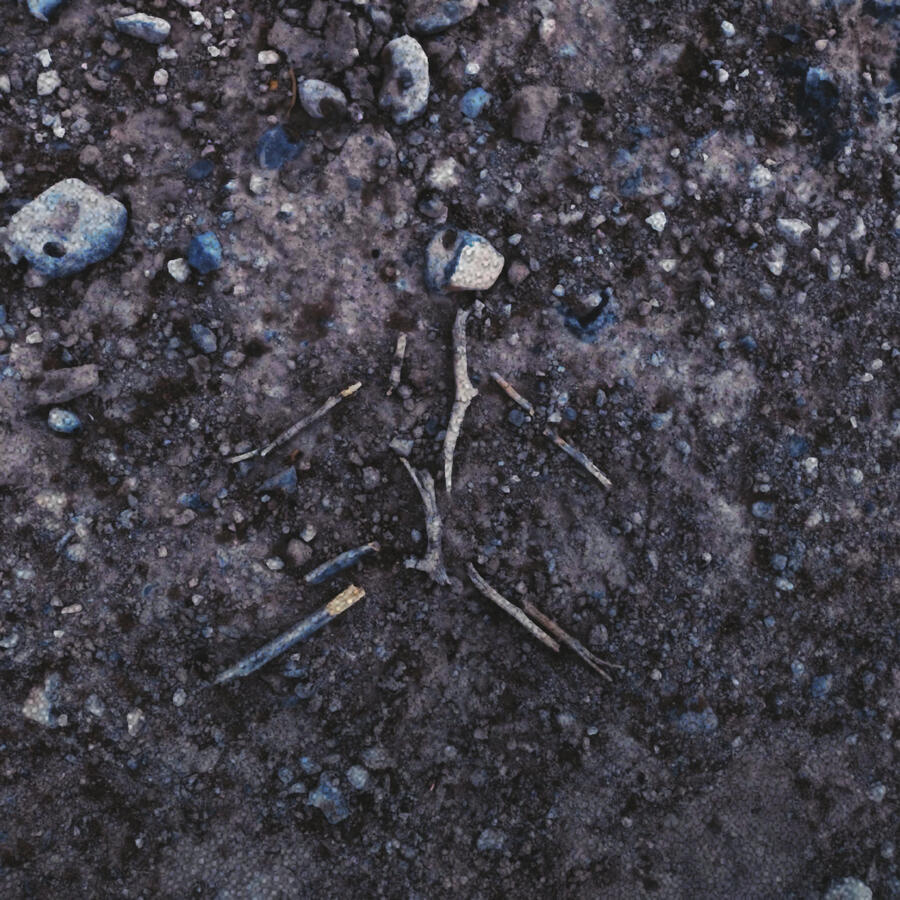 Point of view is facing down the a gravel ground, looking at a stick figure made of sticks and a rock.