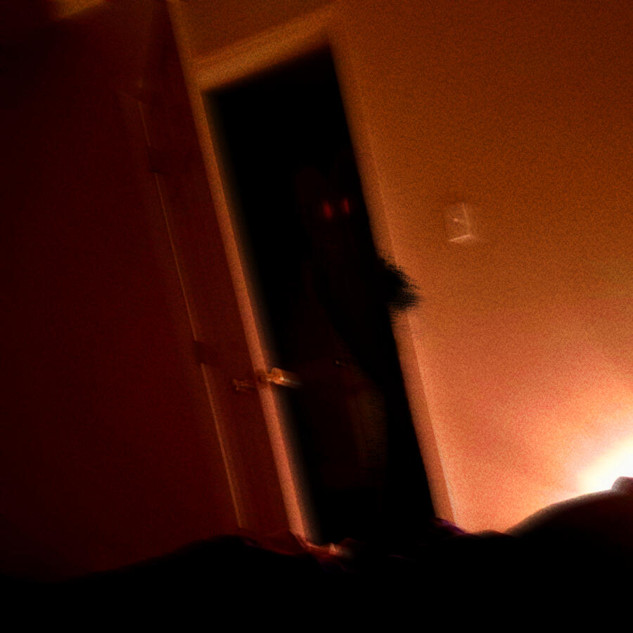 A tilted point of view from a bed, looking through a doorway into the hallway. Standing by the door is a shadow figure with red eyes with a hand peeking in.